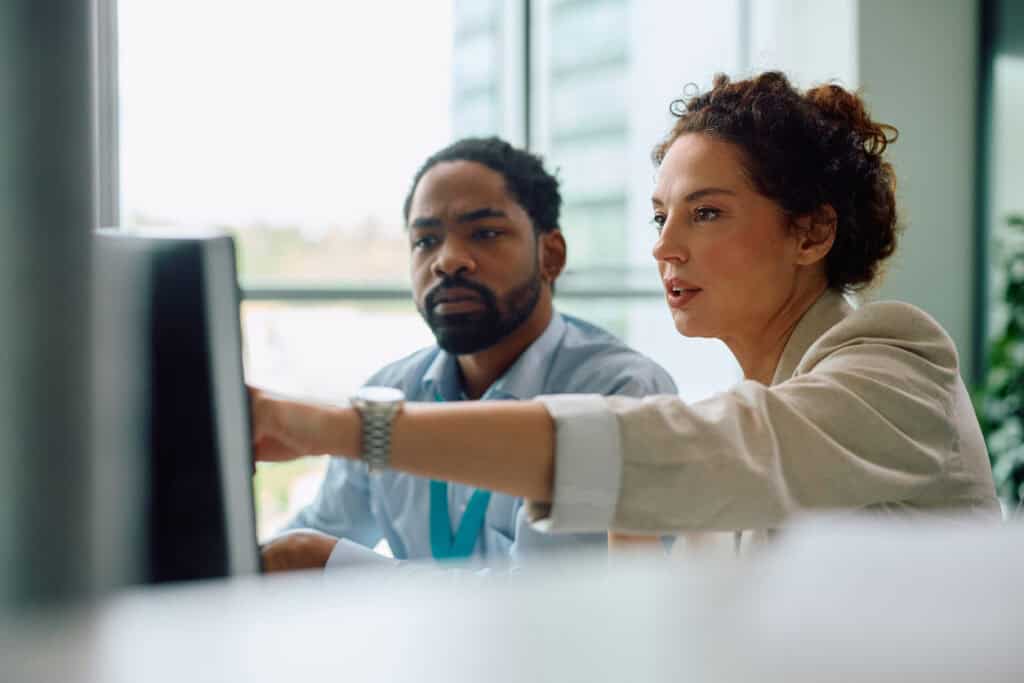 Two business professionals collaborating at a computer, with a woman in a beige blazer pointing at the screen while a man in blue attentively listens.